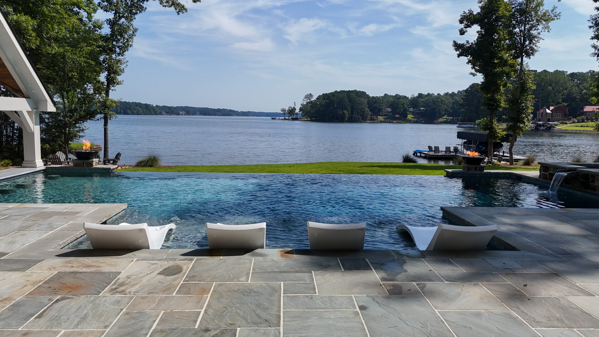 Infinity pool with four white lounge chairs overlooks a serene lake, surrounded by trees and houses. The sky is partly cloudy, creating a peaceful and inviting outdoor scene.