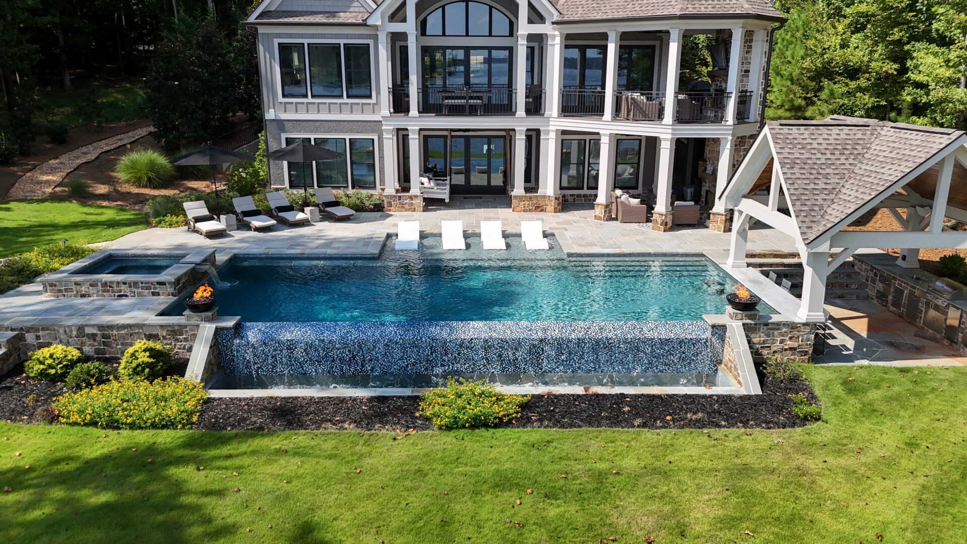 A spacious modern house with an infinity pool that has a waterfall edge, surrounded by sun loungers, a covered patio area, and vibrant green landscaping.