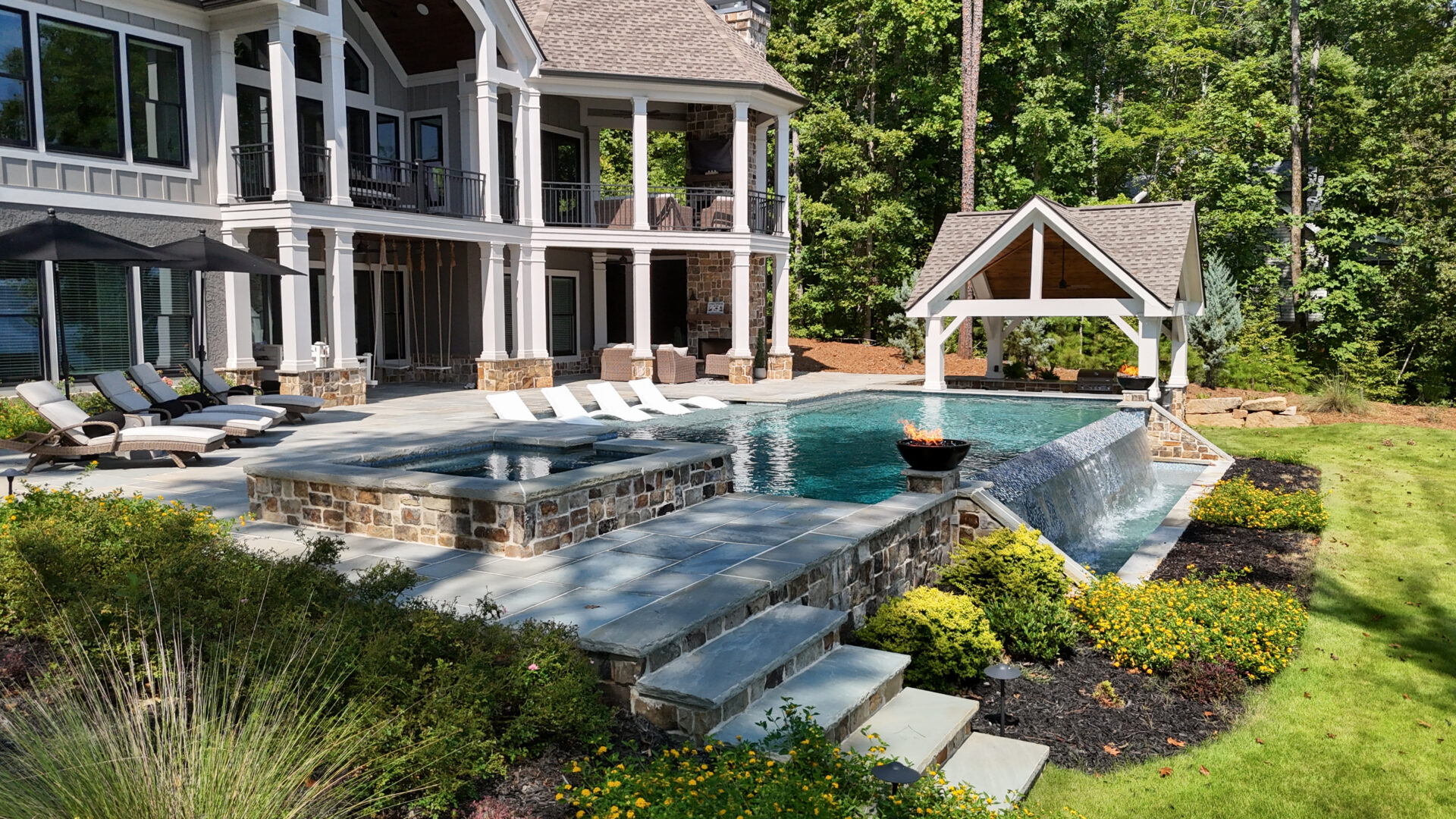 Spacious luxury backyard featuring a pool, hot tub, lounge chairs, and a pavilion, all set within a beautifully landscaped garden next to a large two-story house with stone and wood accents, surrounded by mature trees.