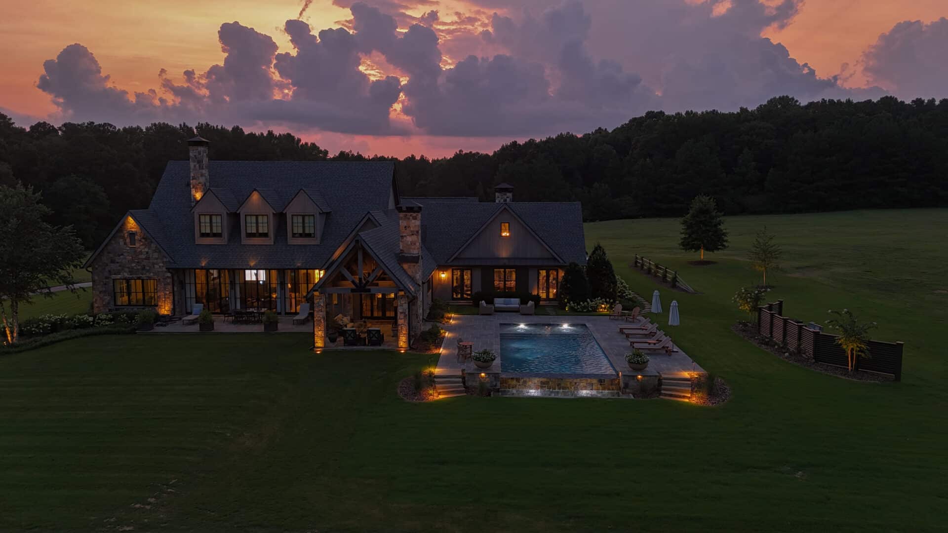 A large house with a pool is illuminated at dusk, set amidst an expansive green lawn and trees, with a vivid sunset sky in the background.