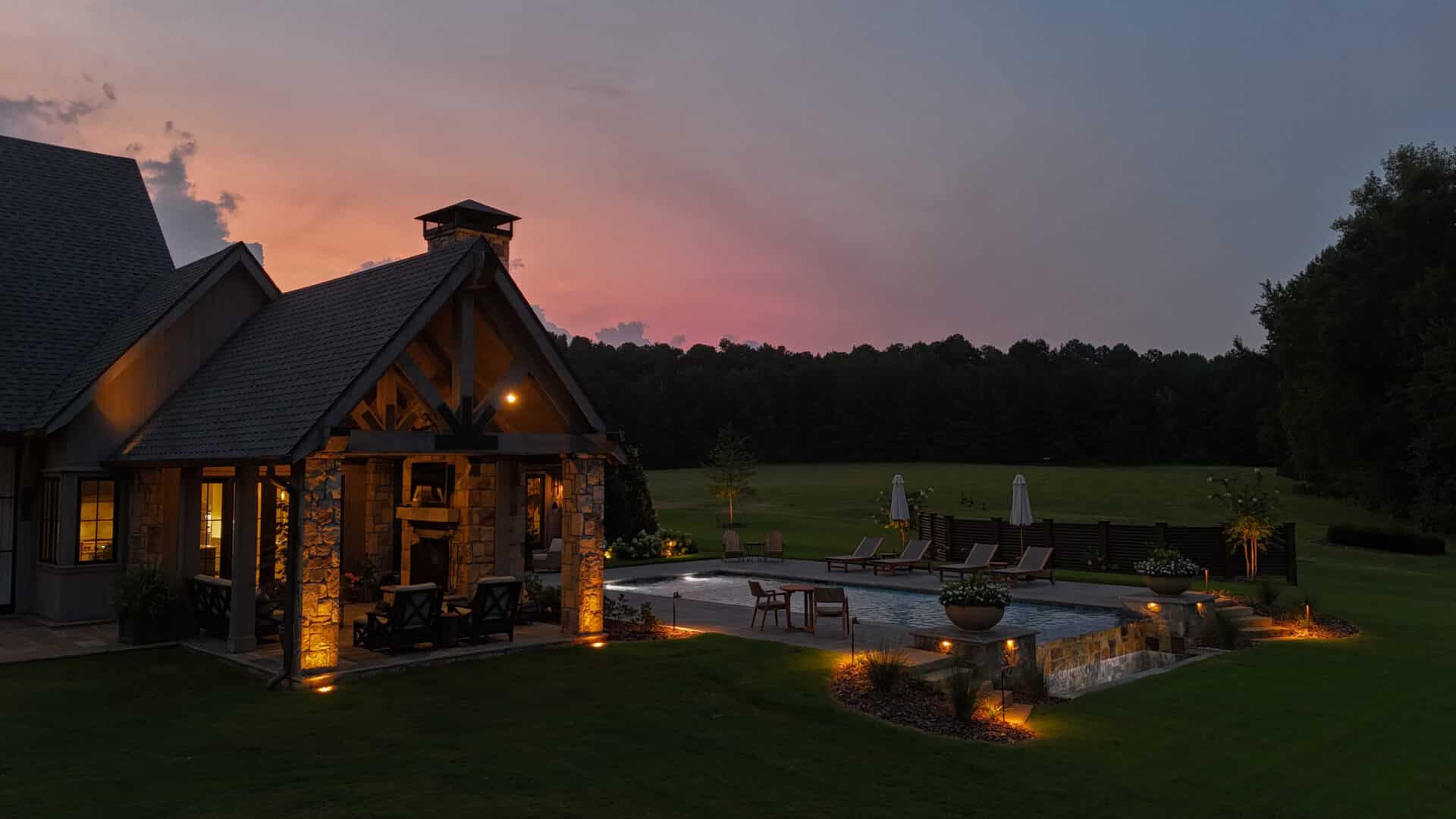 A backyard featuring a pool, lounge chairs, and a brightly lit stone patio next to a house at dusk, bordered by green lawn and trees beneath a vibrant evening sky.