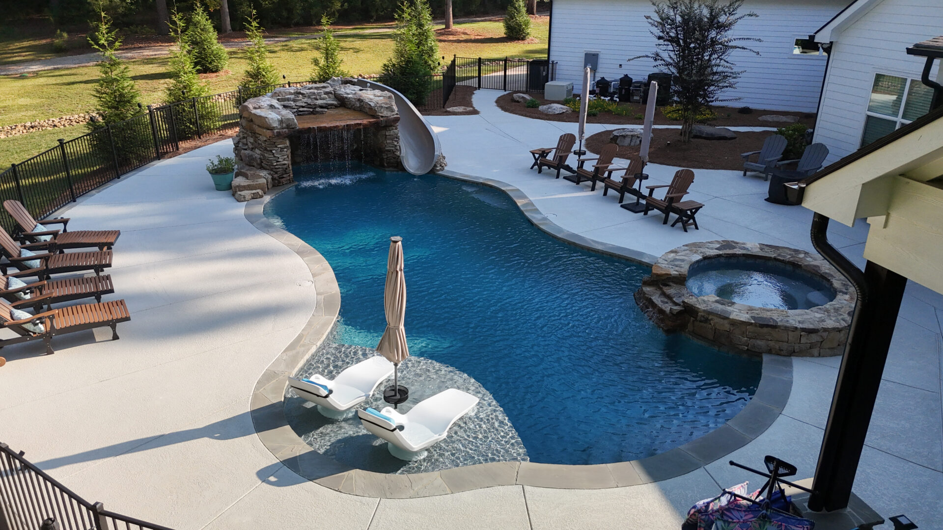 A backyard pool features a rock waterfall slide, a hot tub, and several lounge chairs. Two loungers with umbrellas rest on a shallow ledge in the water. The patio offers plenty of seating, and trees provide a natural backdrop.