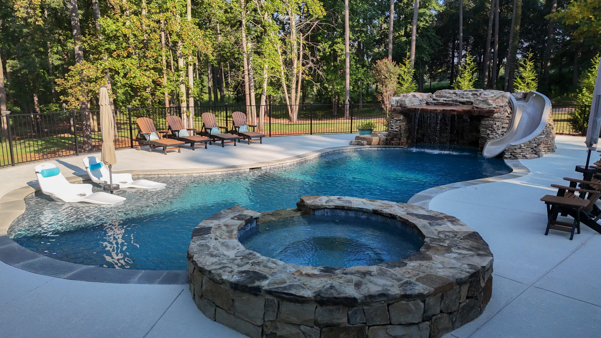 A backyard pool featuring a stone hot tub, waterslide, and waterfall accent, with lounge chairs arranged nearby and trees in the background, all surrounded by a black metal fence.