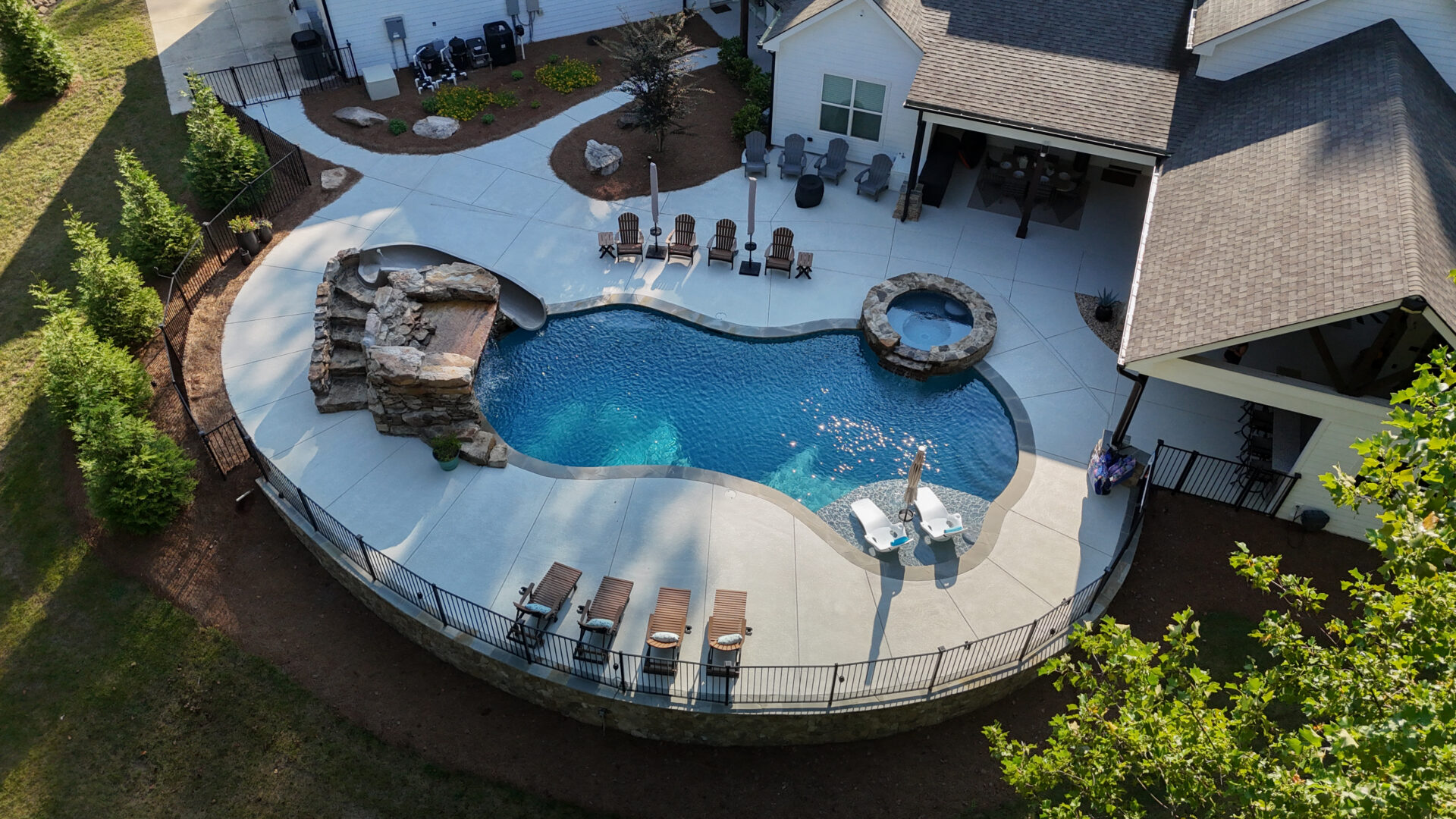 Aerial view of a backyard pool featuring a waterfall, adjacent hot tub, lounge chairs with umbrellas, and a patio area alongside a house, all surrounded by landscaping and enclosed by a black metal fence.
