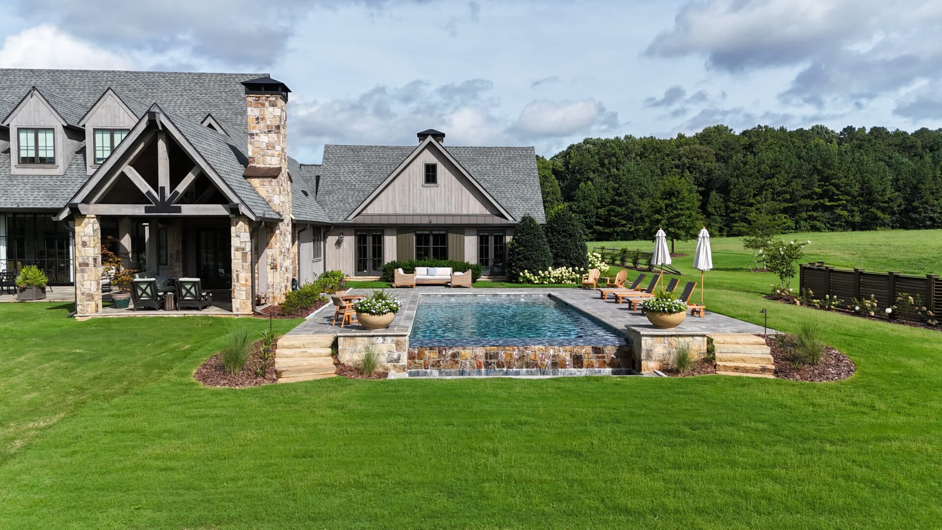 A contemporary farmhouse with stone details overlooks a backyard pool area, complete with lounge chairs and umbrellas, all set amid a well-manicured green lawn and trees beneath a partly cloudy sky.