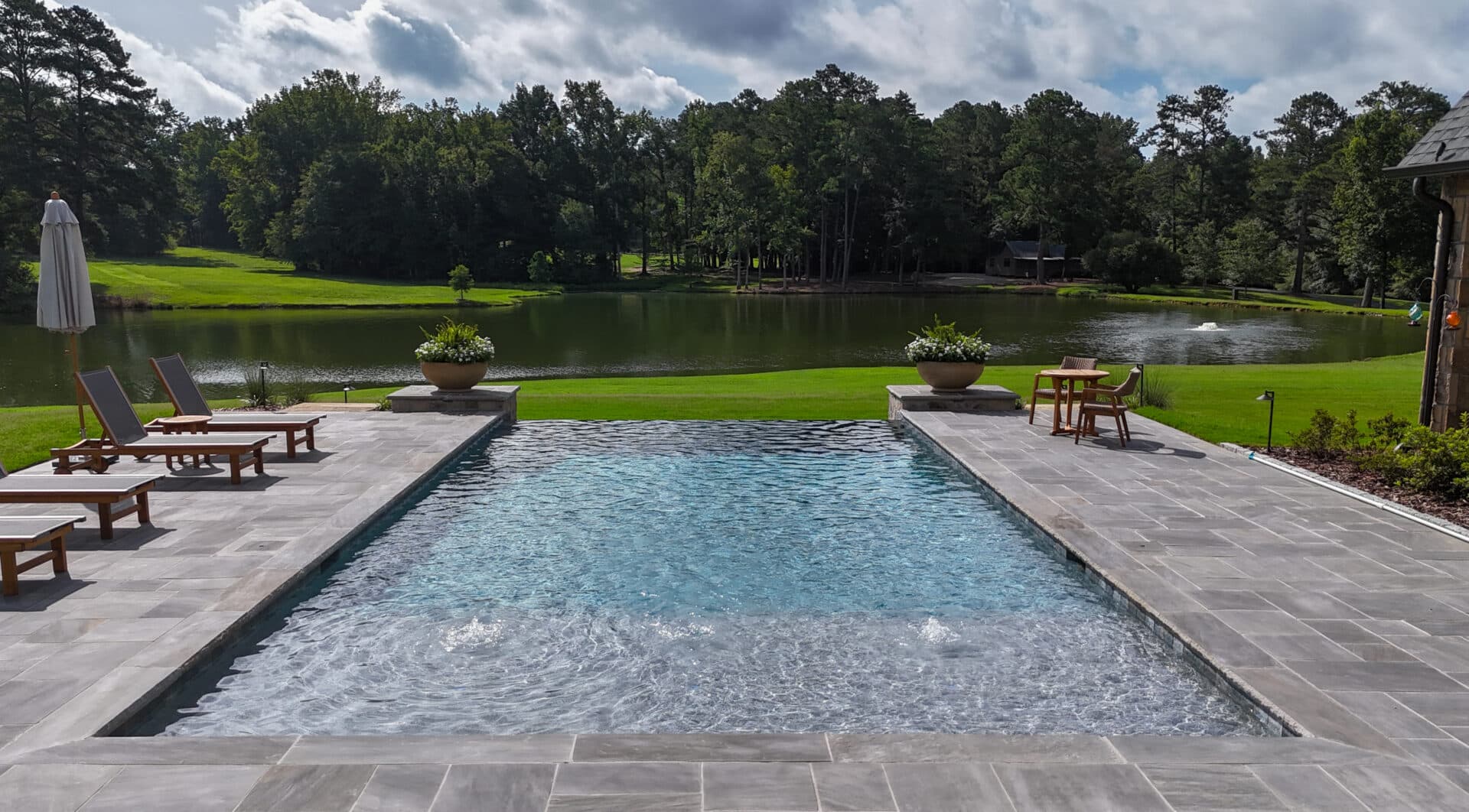 A rectangular pool with lounge chairs on both sides overlooks a green lawn and a serene lake with trees in the background beneath a partly cloudy sky.