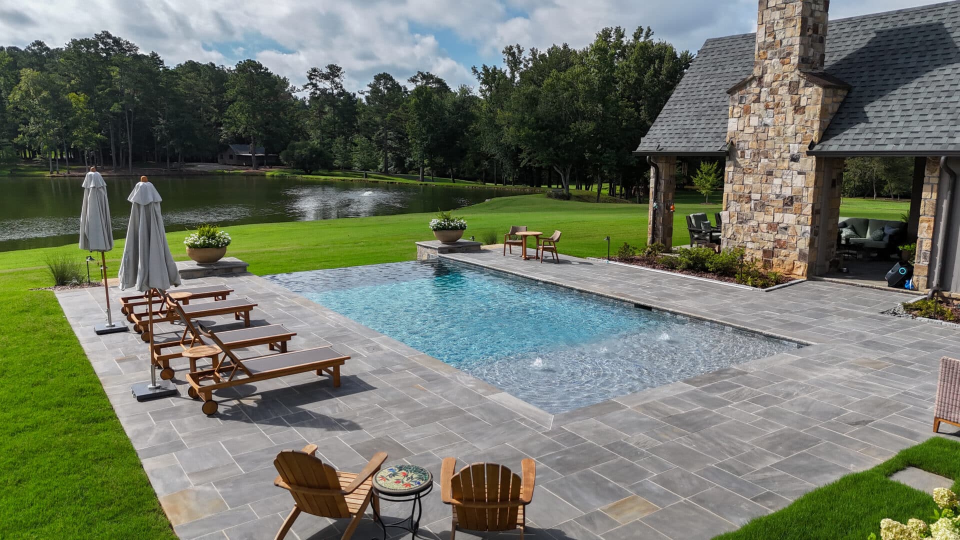 A backyard pool featuring wooden loungers and umbrellas sits on a stone patio, accompanied by a fireplace near a grassy lawn and lake. The area is surrounded by trees beneath a partly cloudy sky.