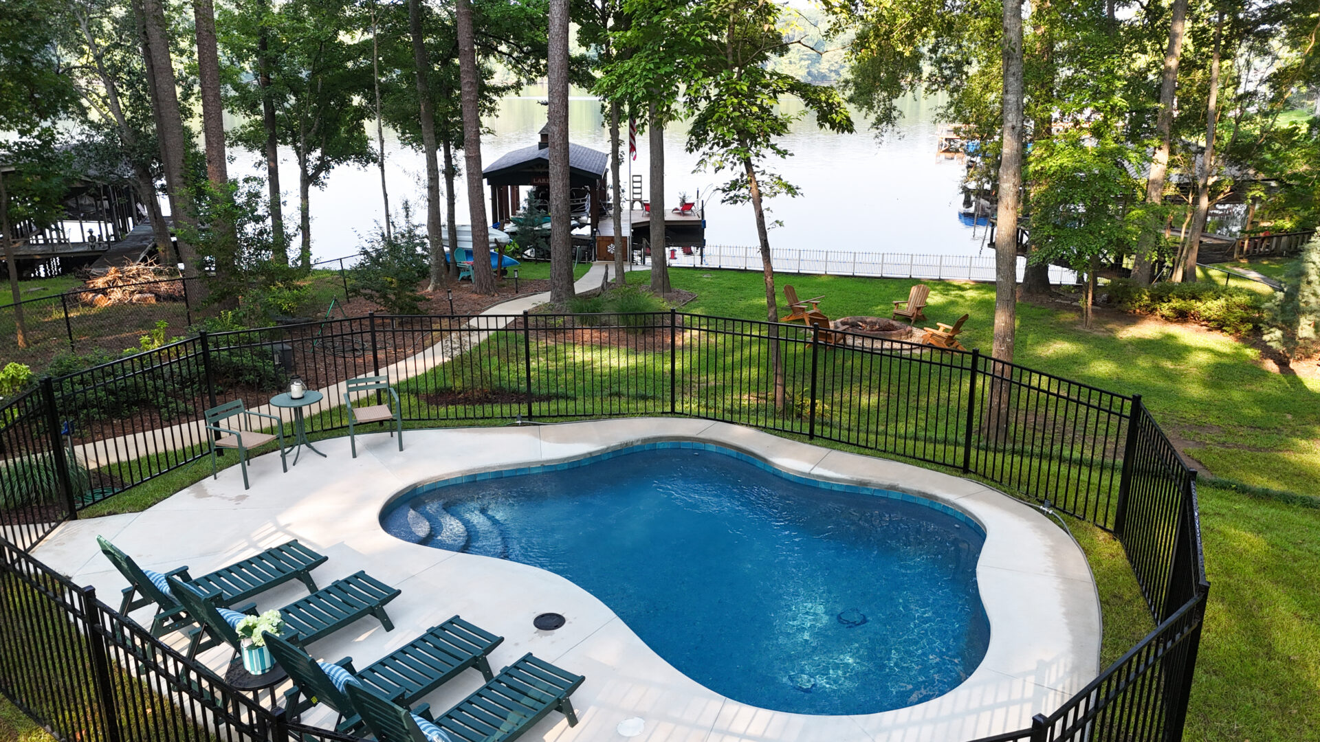 A fenced backyard pool with lounge chairs faces a lake, bordered by trees and lush green grass. Near the water, there's a fire pit area with Adirondack chairs, and a dock extends out into the lake.