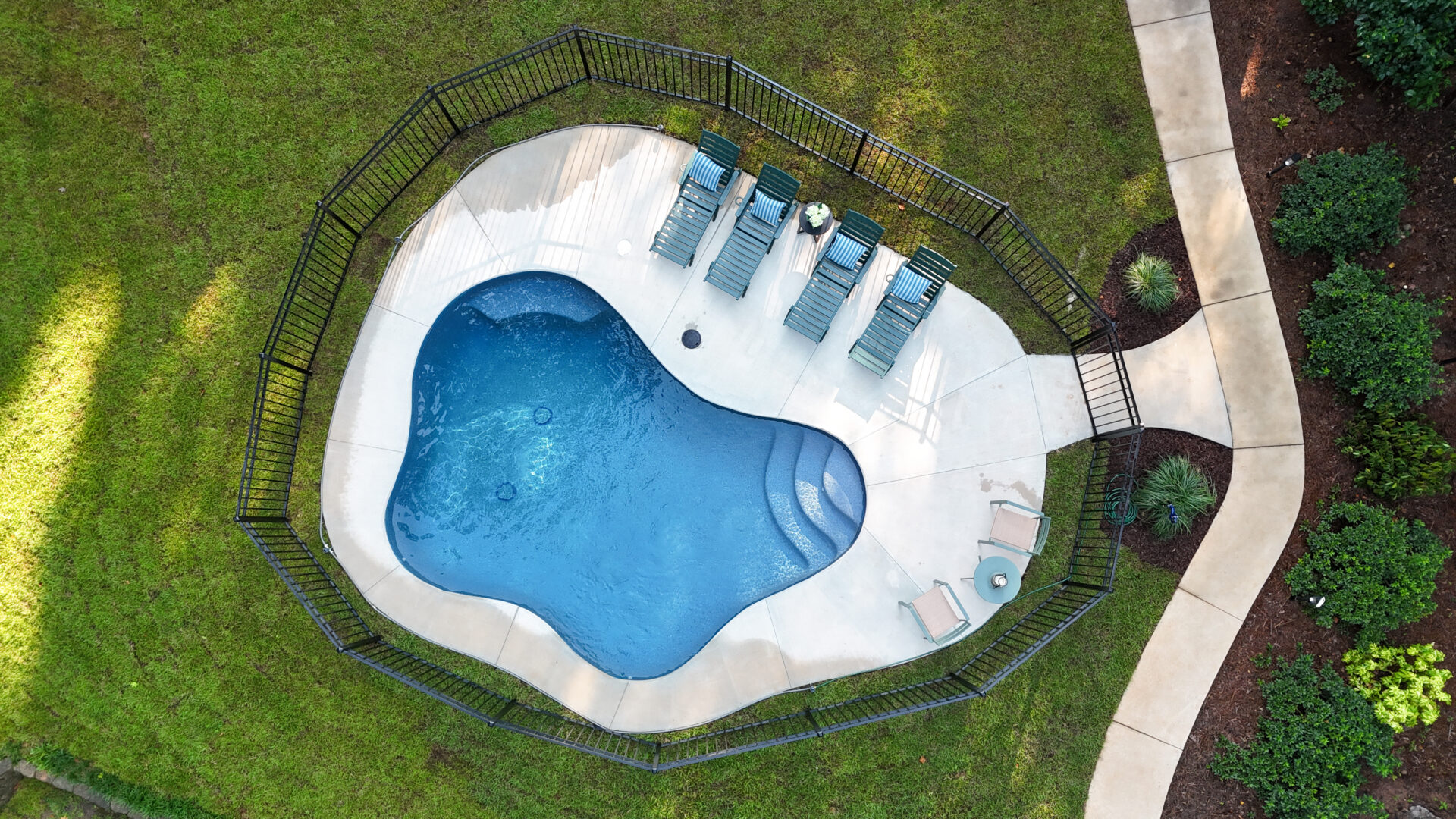 Aerial view of a small kidney-shaped swimming pool with six lounge chairs and two side chairs on a concrete patio, surrounded by grass, landscaping, and a black metal fence.