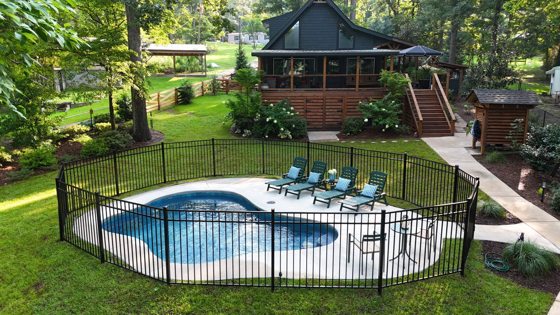 A small fenced-in pool with six lounge chairs is set on a grassy lawn beside a wooden house, surrounded by trees and lush landscaping. A concrete path connects the deck to the pool area.