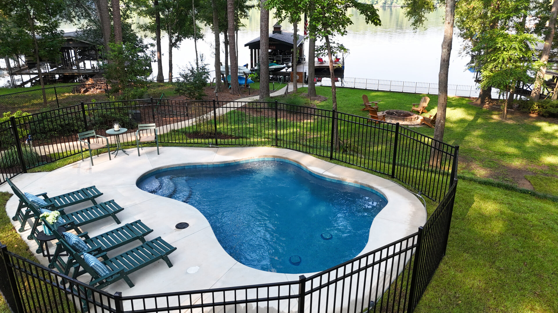 A small fenced backyard pool with lounge chairs beside it, surrounded by green grass and trees. A fire pit with chairs sits near a lakefront dock in the background.