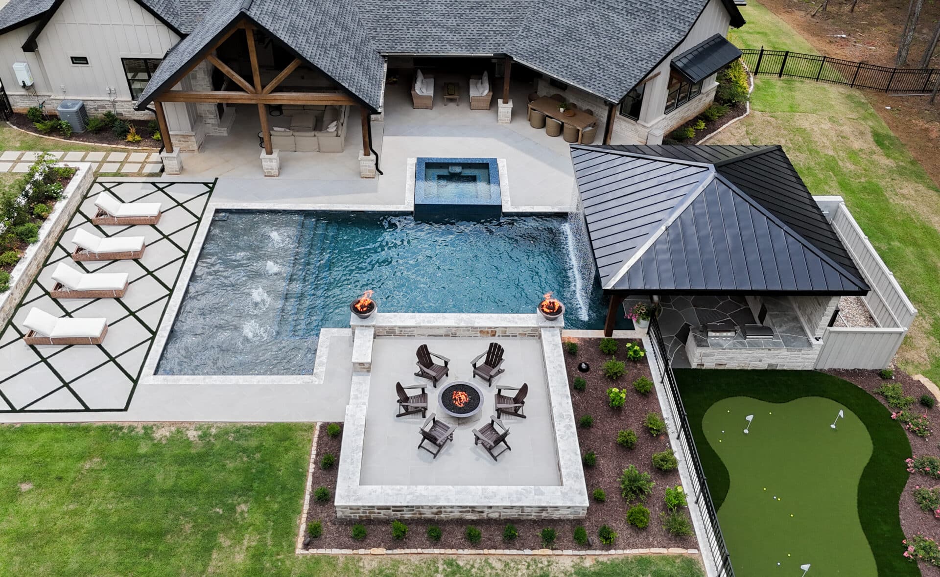 Aerial view of a modern backyard featuring a pool, hot tub, fire pit seating area, covered patio, lounge chairs, and a small putting green surrounded by landscaped grass and plants.