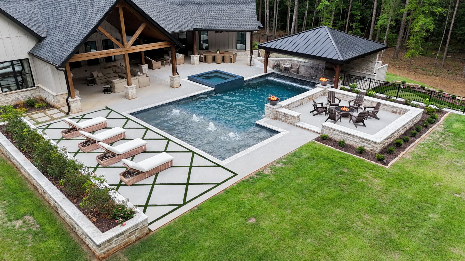 A modern backyard featuring a swimming pool, hot tub, lounge chairs, fire pit seating area, covered patio, and landscaped greenery, all surrounded by trees.