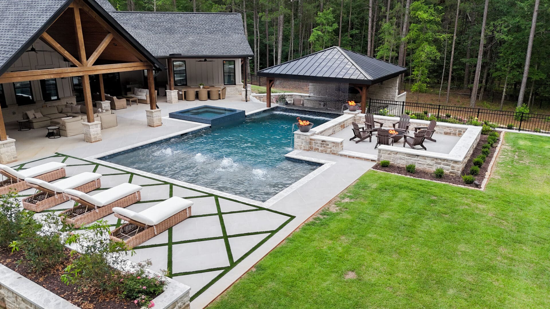 A modern backyard featuring a pool, spa, lounge chairs, an outdoor kitchen, fire pit seating area, and a grassy lawn bordered by trees.