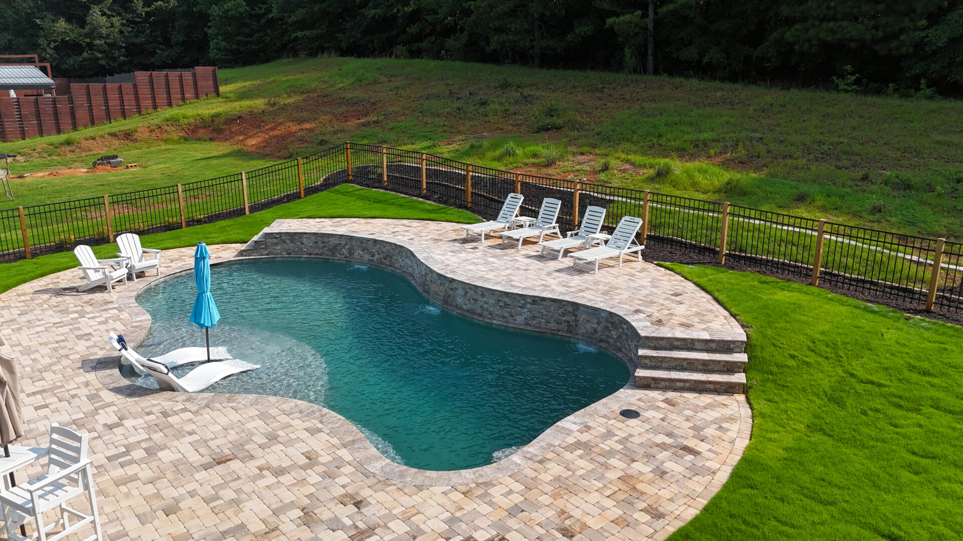 A kidney-shaped backyard pool with a curved stone deck, lounge chairs, and an umbrella. Green grass surrounds the fenced pool area, with a wooded backdrop beyond the fence.