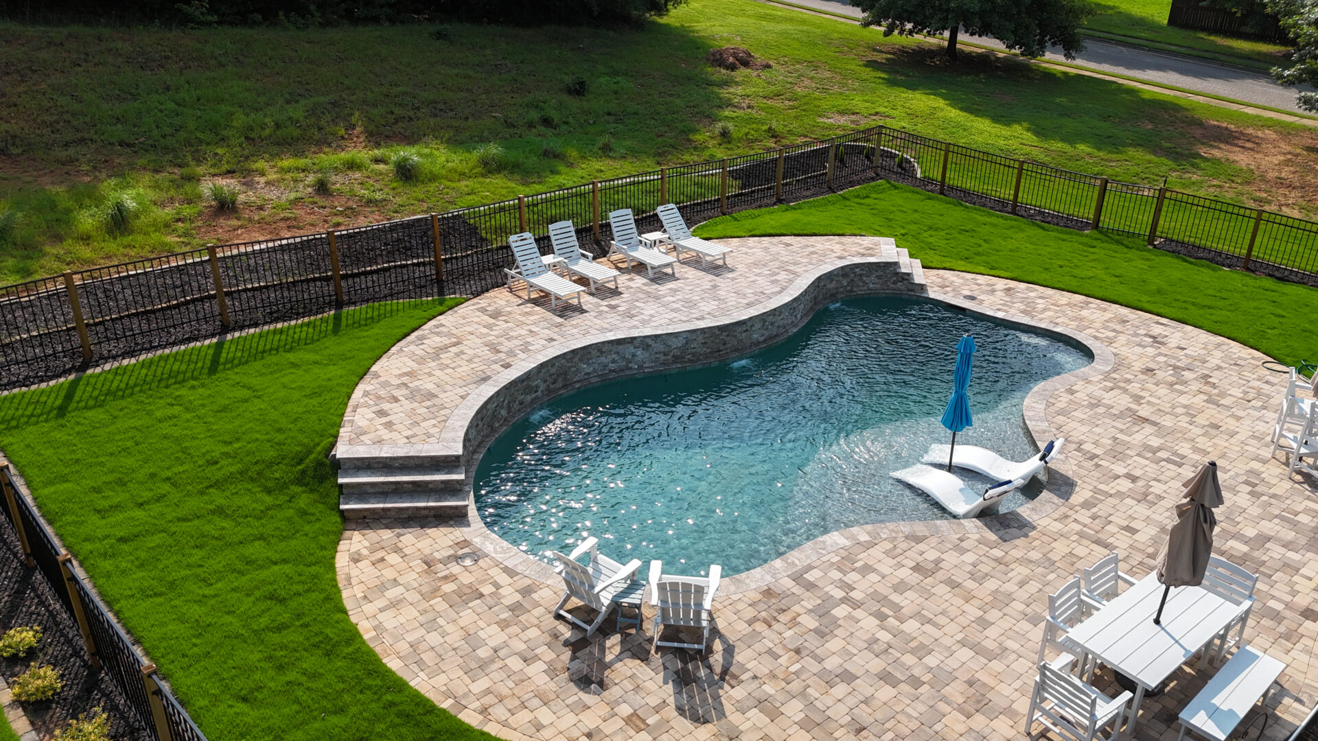 A kidney-shaped backyard pool sits next to a patio with lounge chairs, a blue umbrella, and outdoor dining sets. The fenced pool area is bordered by lush green grass.