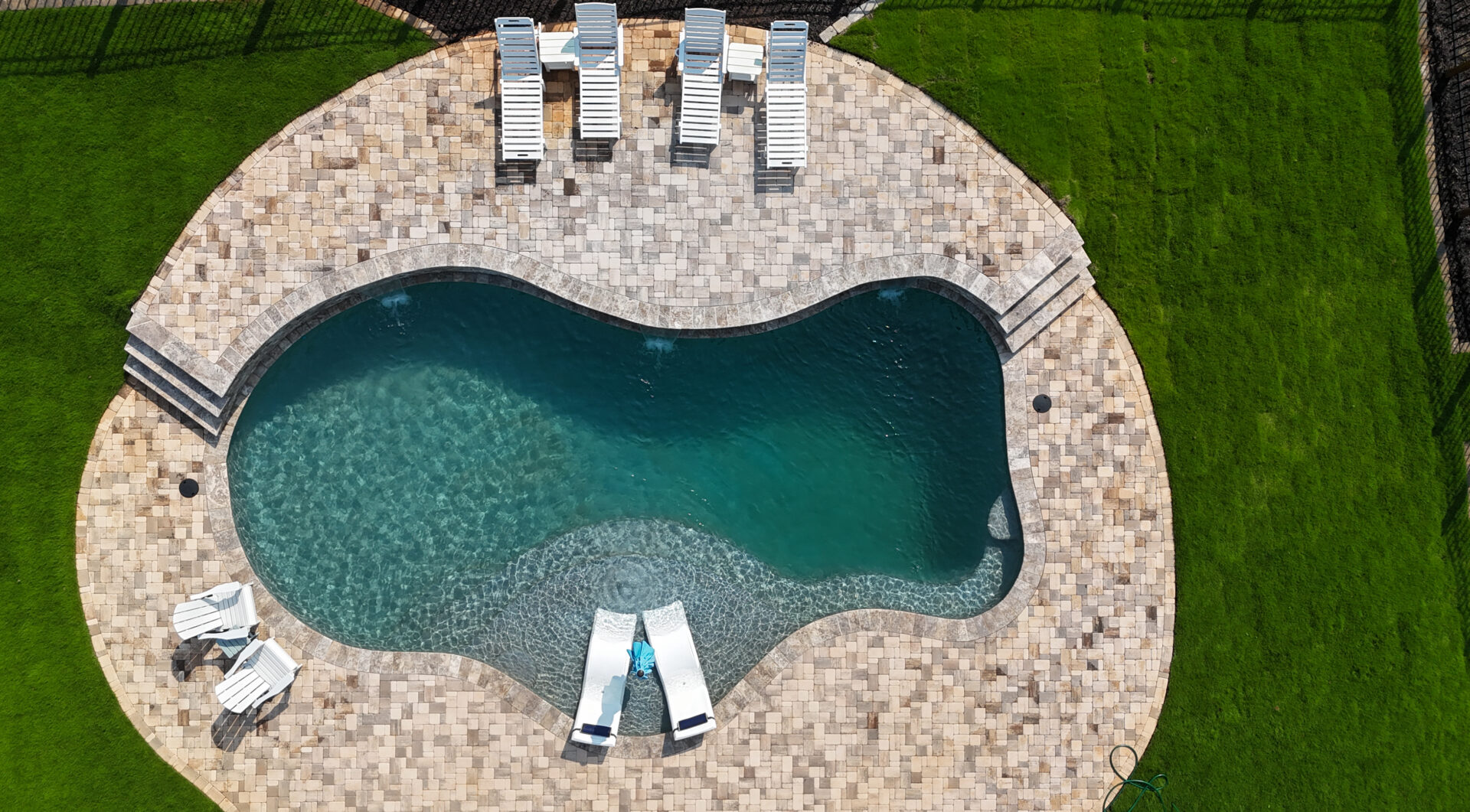 Aerial view of a backyard kidney-shaped pool with clear water, bordered by a stone patio, several lounge chairs, and lush green grass.