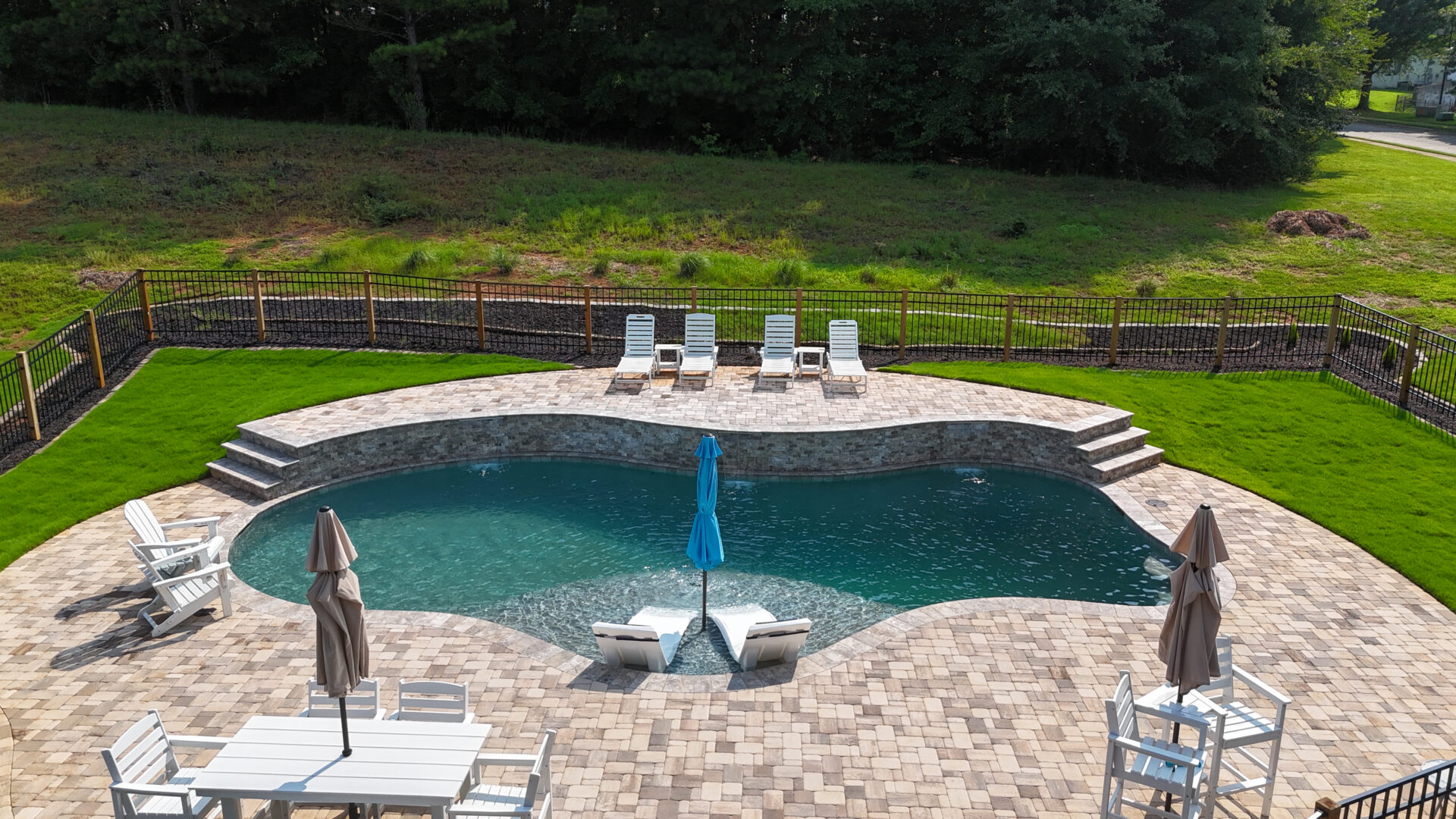 A backyard swimming pool sits on a stone patio furnished with lounge chairs, umbrellas, and tables. The area is enclosed by a black fence and bordered by green grass, with trees visible in the background.