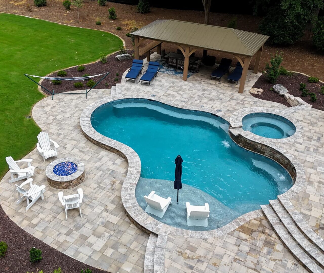Aerial view of a backyard featuring a curvy swimming pool with an attached hot tub, patio chairs arranged nearby, a fire pit, hammock, covered seating area, picnic table, and green lawn surrounding the space.