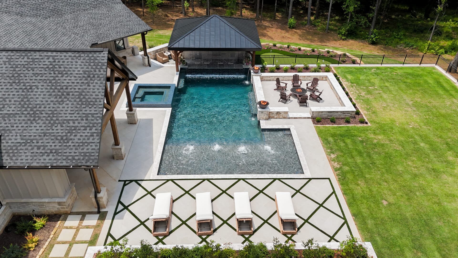Aerial view of a backyard featuring a pool, hot tub, shaded lounge area, fire pit surrounded by chairs, neatly manicured lawn, and contemporary landscaping.