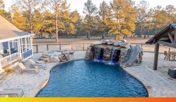 A backyard pool featuring a rock waterfall, a water slide, lounge chairs, and a patio area, all enclosed by a wooden fence and trees beneath a clear sky.