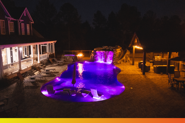 A backyard pool at night glows with purple lighting, showcasing a waterfall, submerged lounge chairs, and a fire pit. The patio area and house nearby are bathed in a warm, inviting light.