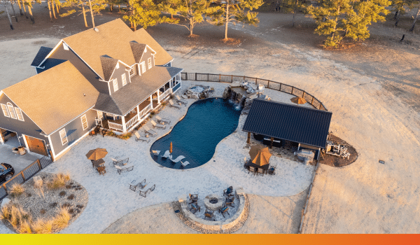 Aerial view of a house featuring a backyard pool, poolside building, lounge chairs, umbrellas, and a fire pit, all surrounded by trees and sandy terrain.