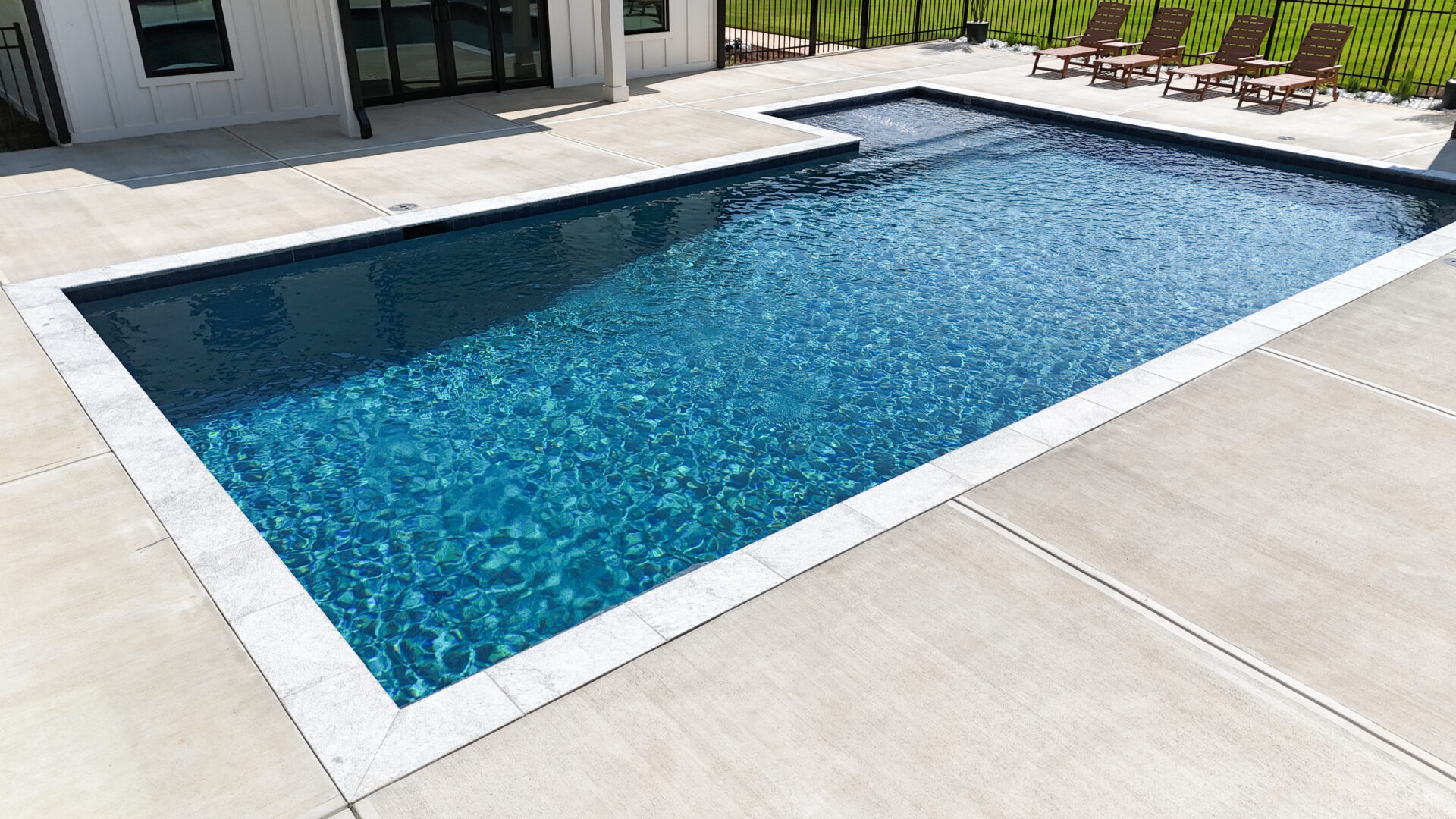 A rectangular outdoor swimming pool with clear blue water is bordered by a light-colored concrete deck. Four brown lounge chairs sit in a row along one side, positioned near a fence and adjacent to a modern building.
