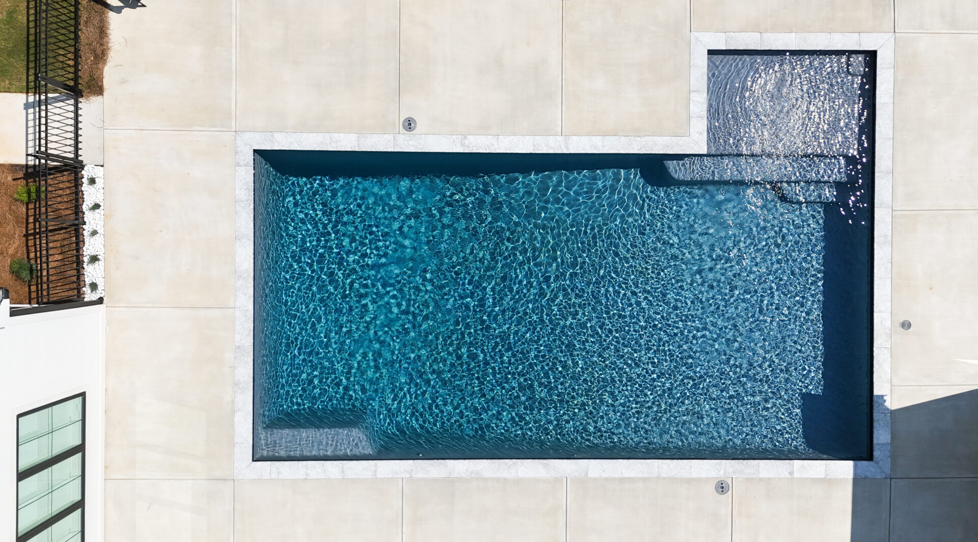 Aerial view of a rectangular swimming pool with clear blue water, surrounded by a light-colored concrete deck. A small attached shallow area is visible in the upper right corner.