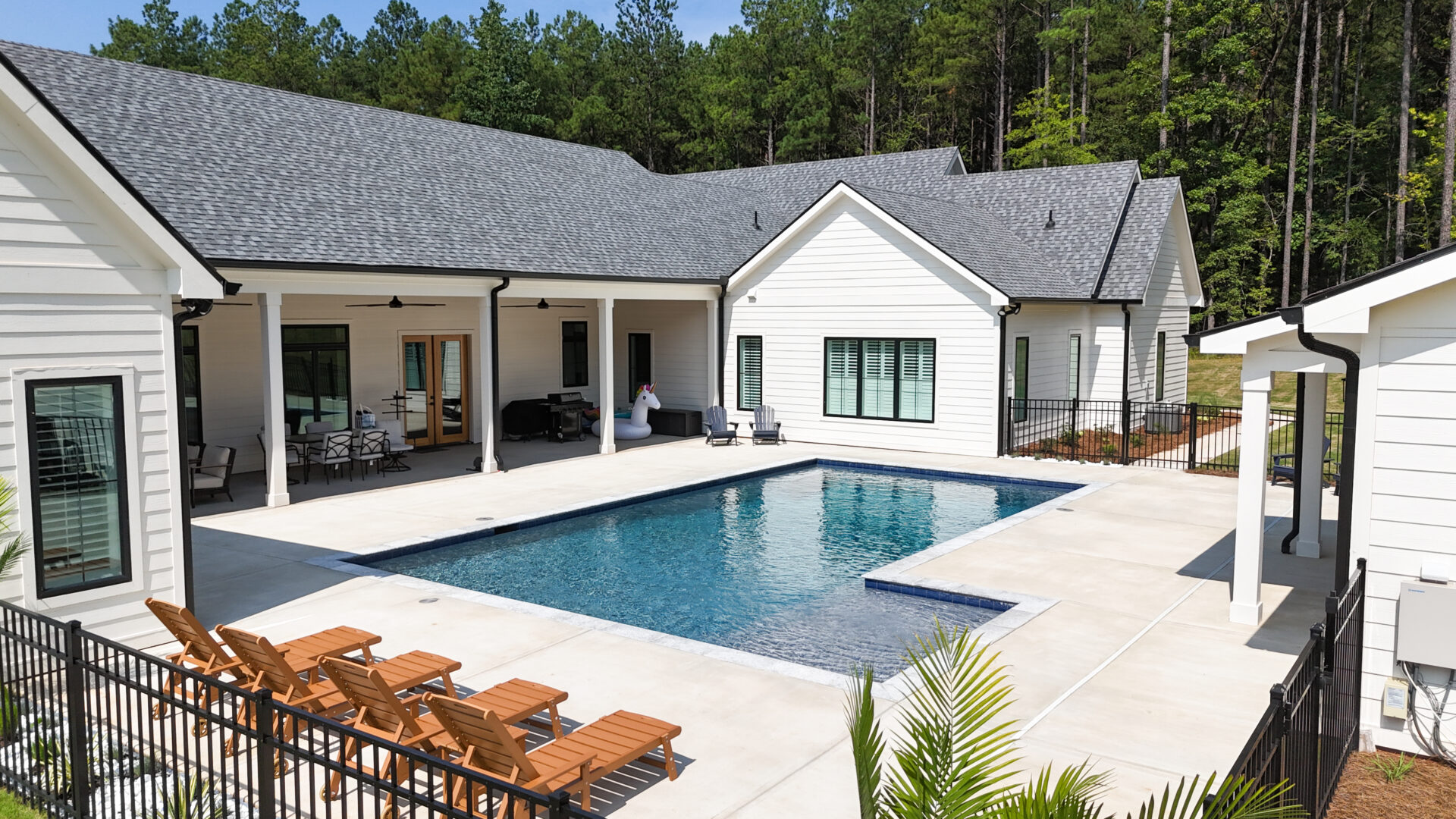 Modern white house with a backyard pool surrounded by trees. Four wooden lounge chairs line the pool deck, and a covered patio with outdoor seating is visible. A person stands near the pool.