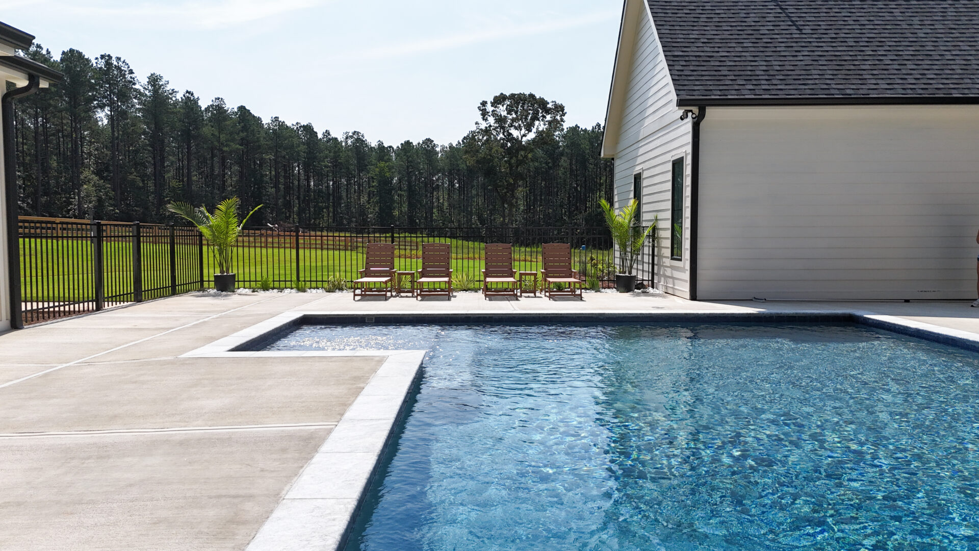 A sparkling outdoor swimming pool sits next to a sleek white modern house. Five brown lounge chairs are arranged neatly along the poolside, accompanied by two potted plants. In the background, there’s a lush grassy yard bordered by tall trees.