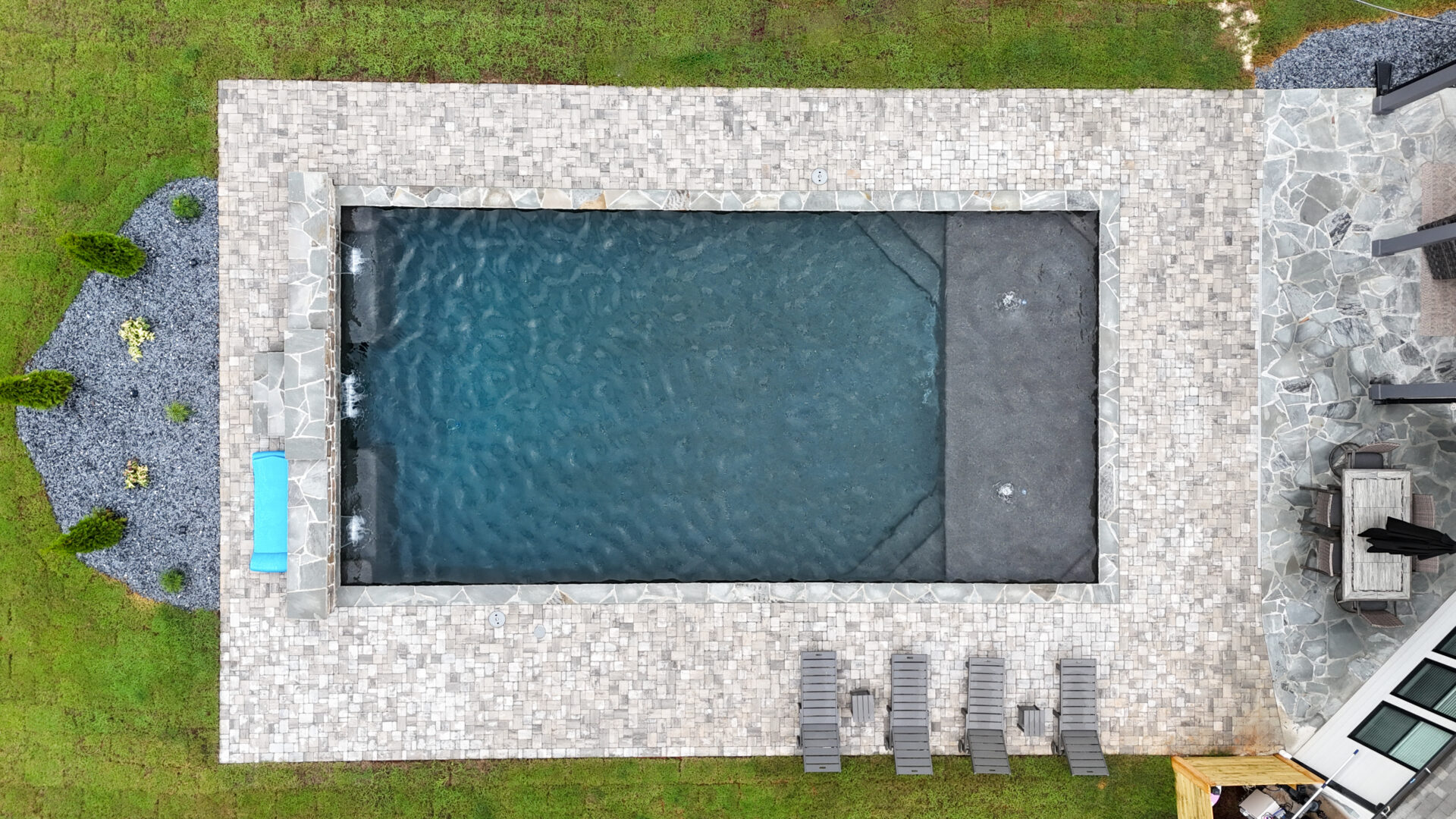 Aerial view of a rectangular backyard pool surrounded by stone tiles, with six lounge chairs lined up on one side and a small water feature on the left. Grass and landscaped areas border the pool.