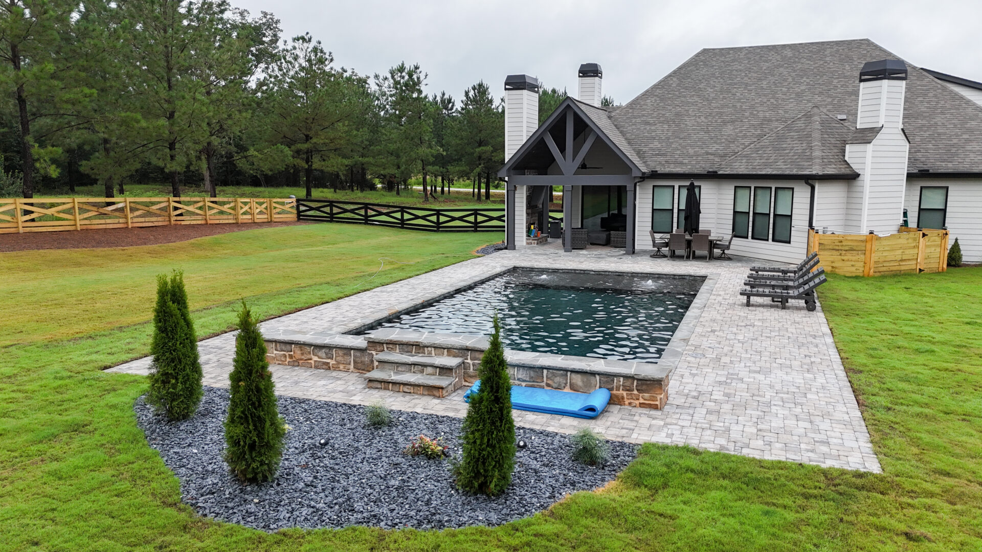 A backyard featuring a rectangular pool and stone patio, surrounded by shrubs and lounge chairs positioned near a modern house. A green lawn and wooden fences enclose the area, with trees visible in the background.