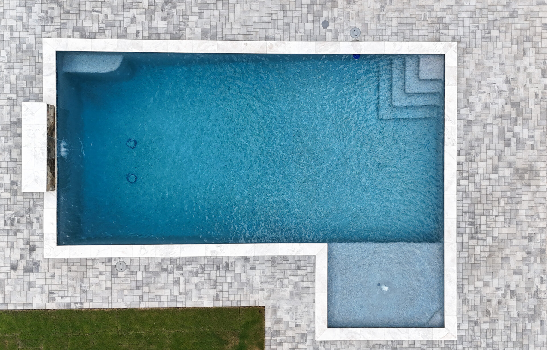A rectangular swimming pool with clear blue water features a shallow ledge on one side and steps in the corner, surrounded by light gray paving stones and a strip of grass in the bottom left corner.