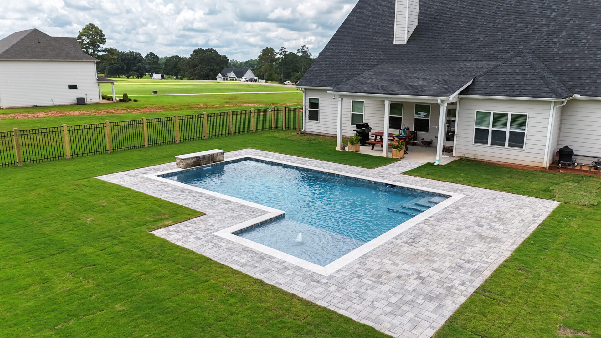 A rectangular backyard pool featuring a small stone waterfall sits within a paved patio and green lawn next to a white house with a spacious covered porch.