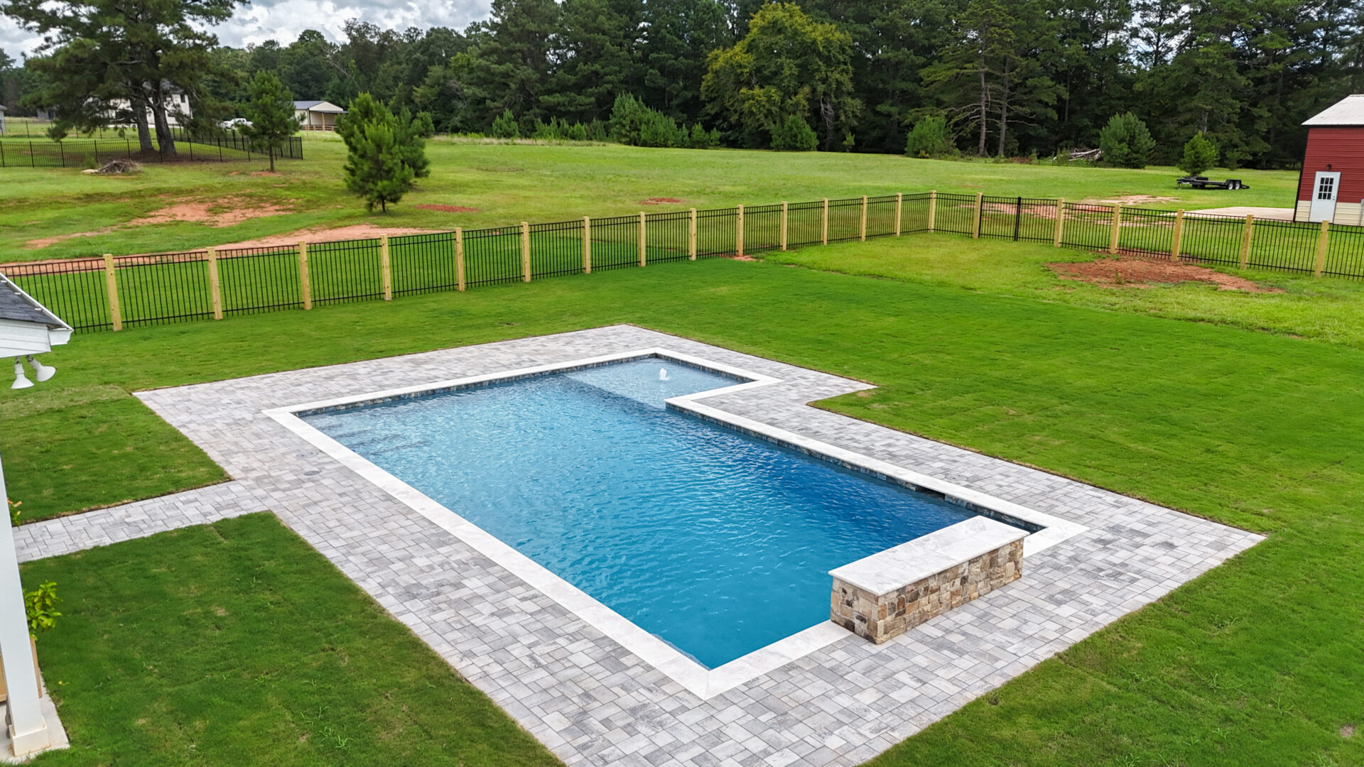 Rectangular backyard pool with gray stone decking, bordered by green grass and a wooden fence, situated in a large open yard with trees in the background.