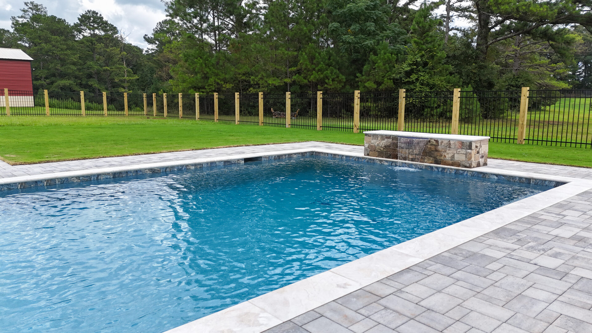 A rectangular backyard pool with clear blue water, stone trim, and a small stone waterfall feature. The pool is surrounded by grass, trees, and a wooden fence.