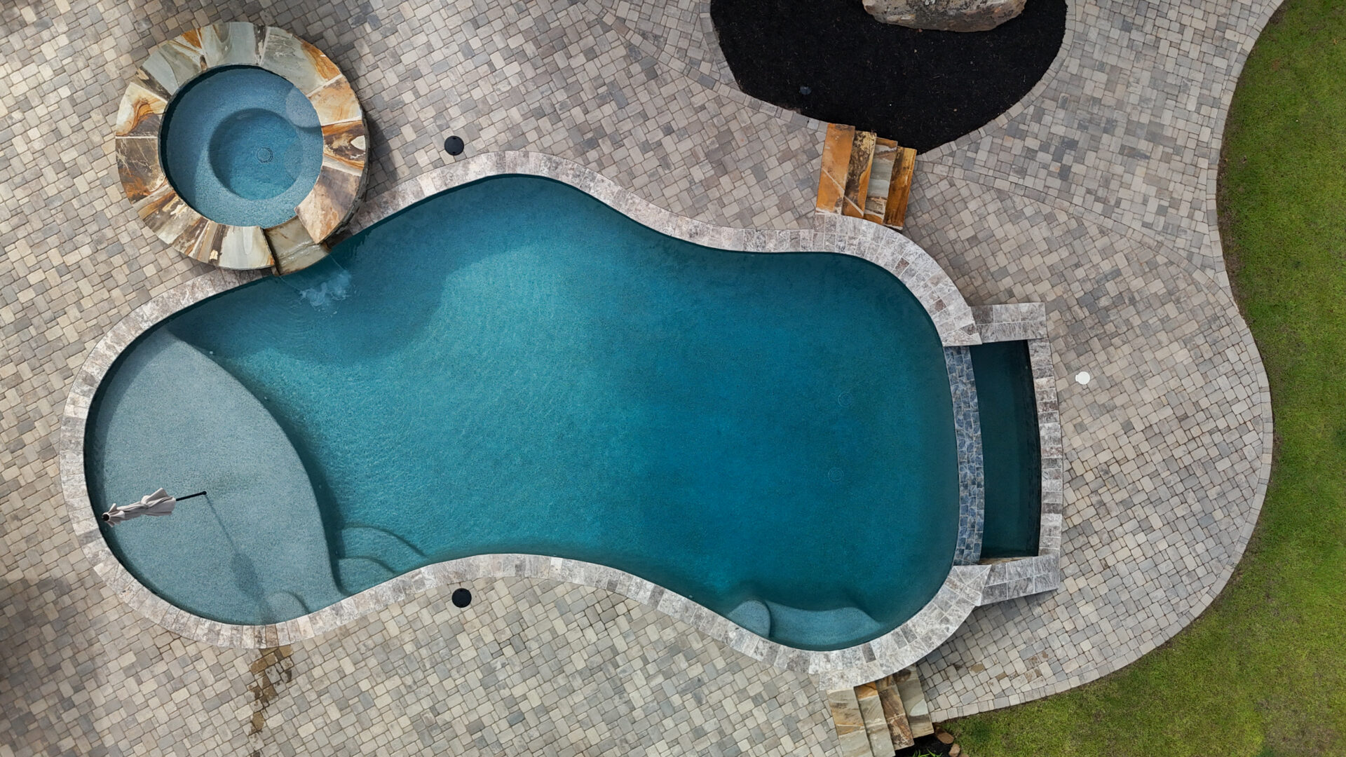 A kidney-shaped swimming pool with an adjacent circular hot tub, surrounded by stone tiles. A person is swimming near the edge, and green grass borders part of the pool area.