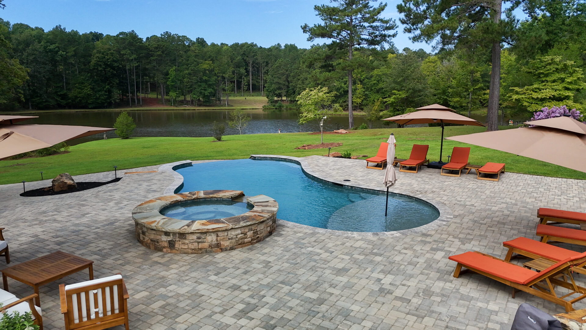 A stone patio featuring a pool and hot tub is surrounded by orange lounge chairs and umbrellas, offering views of a green lawn and a tree-lined lake beneath a clear blue sky.