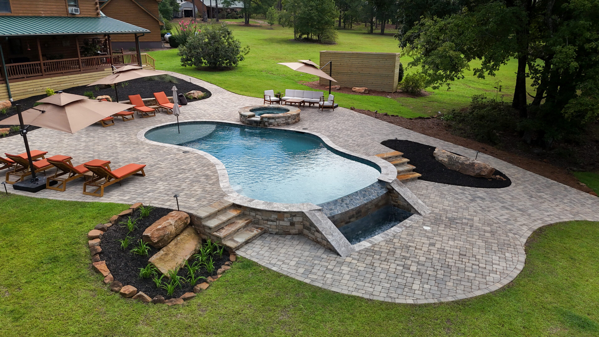 A backyard featuring a freeform pool and an attached hot tub, with patio chairs that have orange cushions, umbrellas providing shade, and landscaped sections. The area is surrounded by lush green grass and trees.