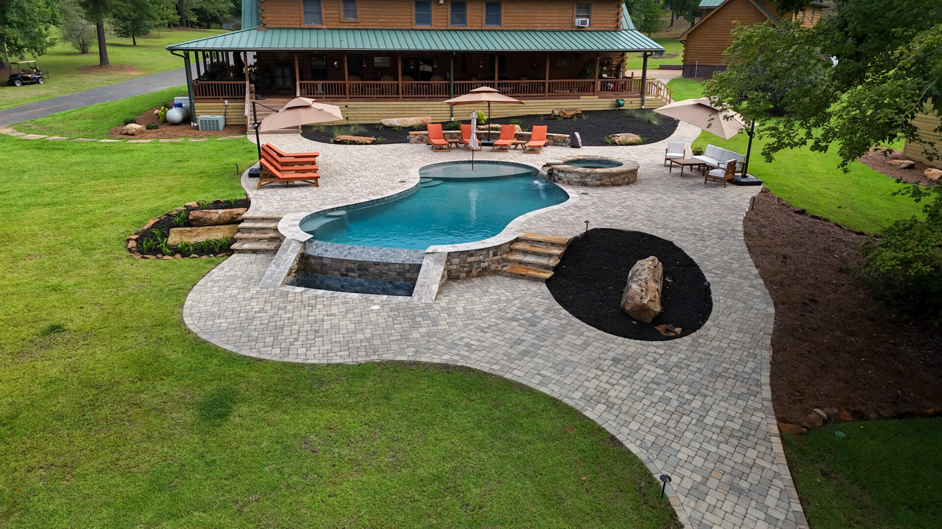 A curved pool featuring an attached hot tub, with stone pavers around it, lounge chairs, umbrellas, and landscaped garden beds in a lush green backyard close to a large wooden house.