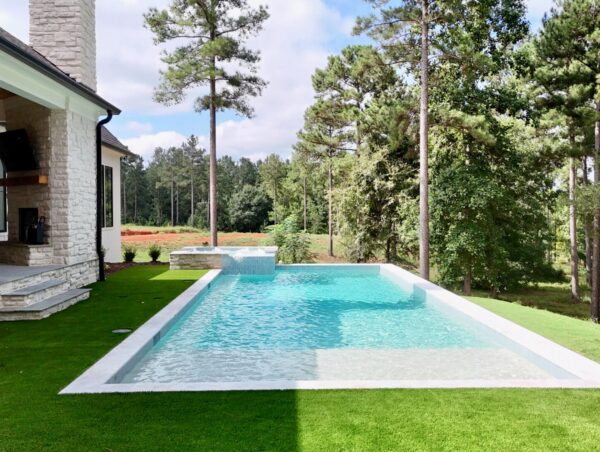 Modern backyard featuring a rectangular infinity pool beside a stone house, bordered by lush artificial grass and tall pine trees, with views of a wooded area beneath a partly cloudy sky.