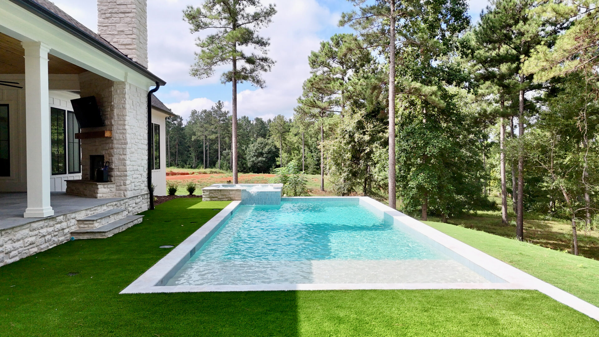Modern backyard featuring a rectangular infinity pool beside a stone house, bordered by lush artificial grass and tall pine trees, with views of a wooded area beneath a partly cloudy sky.