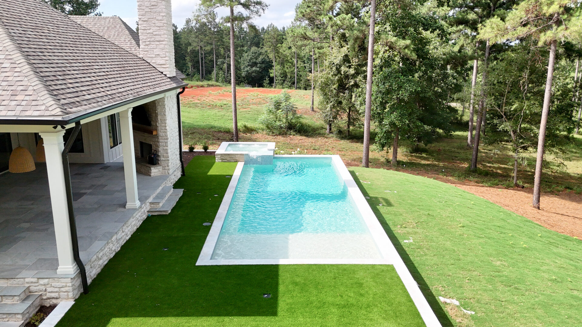 A modern backyard features a rectangular swimming pool with an attached spa, bordered by lush green grass and tall trees. A stone house with a covered patio sits nearby.