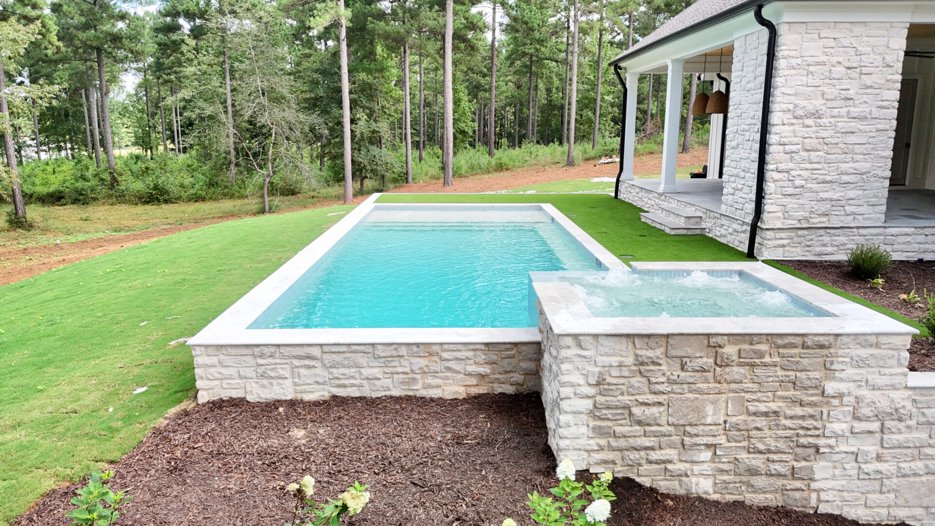 A rectangular swimming pool with an attached hot tub is next to a white brick house, surrounded by a grassy yard and bordered by trees.