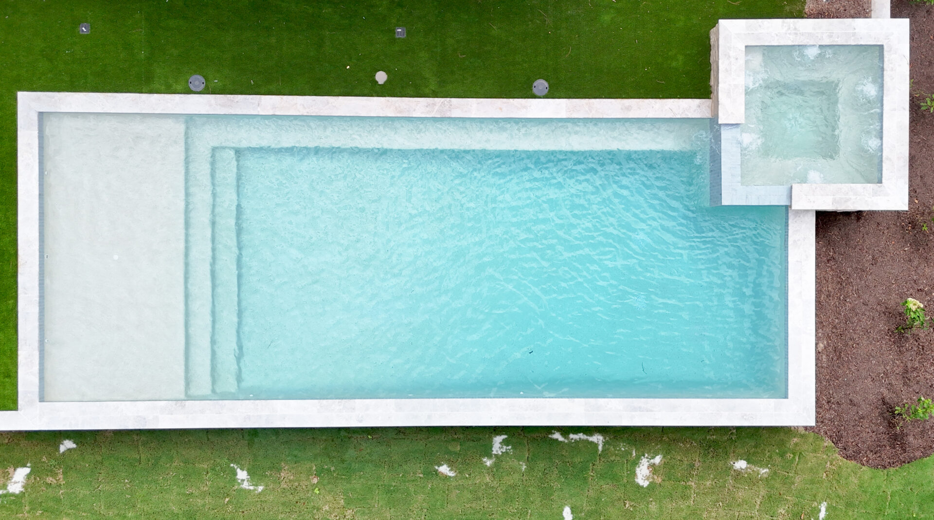A rectangular swimming pool with clear blue water and an attached square hot tub, surrounded by green grass. A few scattered patches of dirt are visible on the grass.