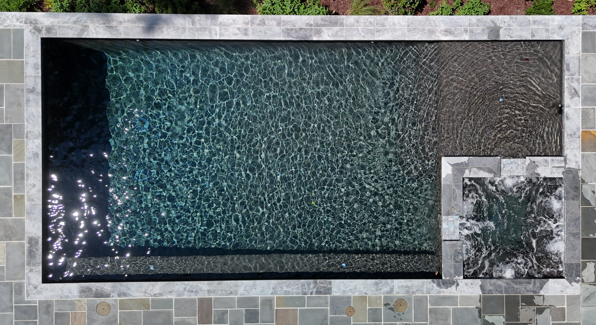 A rectangular swimming pool with clear, rippling water, viewed from above. There is a small, attached square hot tub in the corner and gray stone tiles surround the pool. Some greenery is visible at the top of the image.