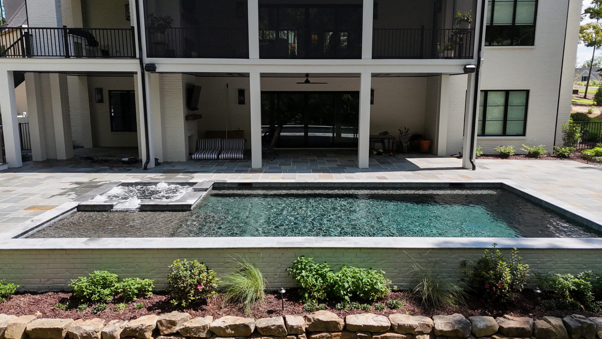 Rectangular backyard pool with a small spa, surrounded by stone tiles, bordered by a low stone wall and landscaping. The pool is in front of a modern two-story house with large windows, balcony, and covered patio.