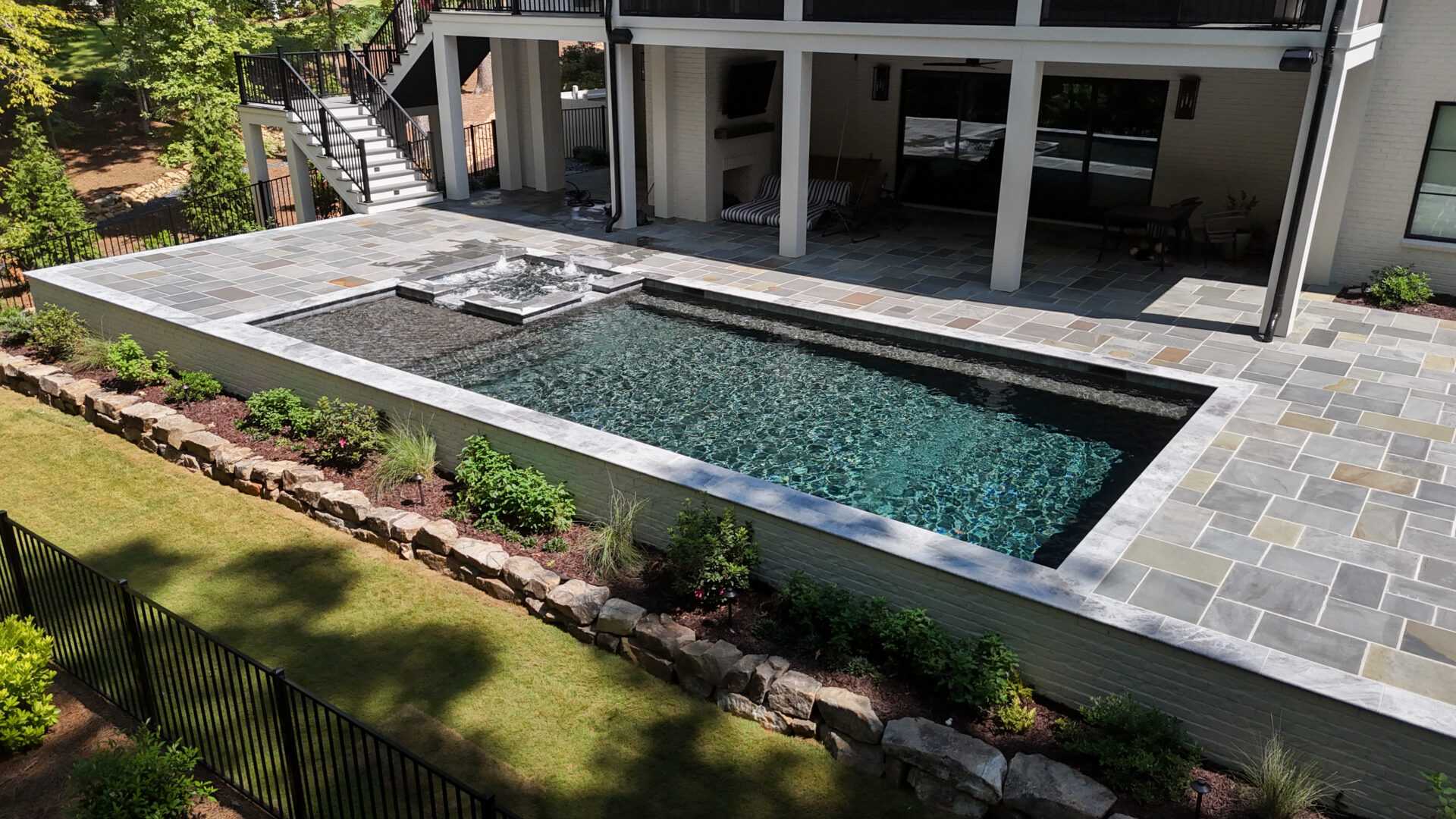Rectangular backyard pool featuring a raised spa, bordered by stone pavers and landscaped with small plants. A fence encloses the yard, with grassy areas and a rock border. The pool sits adjacent to a modern patio area and a white house.