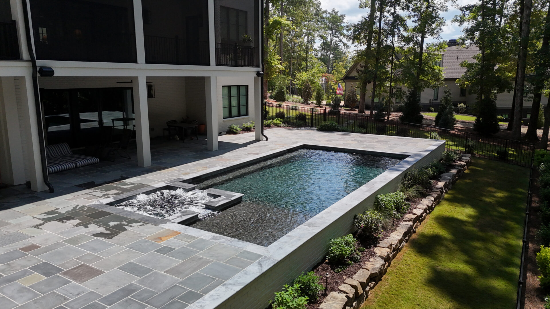 Modern backyard featuring a rectangular pool and hot tub, surrounded by a stone patio and landscaped greenery, next to a house and lush green lawn, with trees and other homes visible in the background.